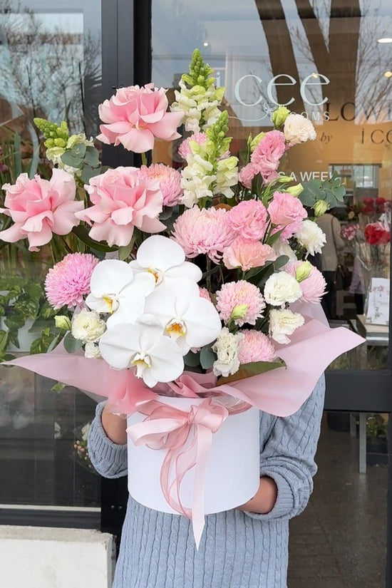Person holding large luxury flower arrangement in white presentation box with pink ribbon, featuring white orchids, pink roses, pink chrysanthemums, and white snapdragons, displayed outside i.ceé Blooms storefront in St Albans