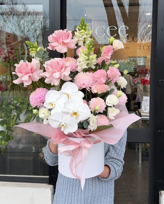 Person holding large luxury flower arrangement in white presentation box with pink ribbon, featuring white orchids, pink roses, pink chrysanthemums, and white snapdragons, displayed outside i.ceé Blooms storefront in St Albans